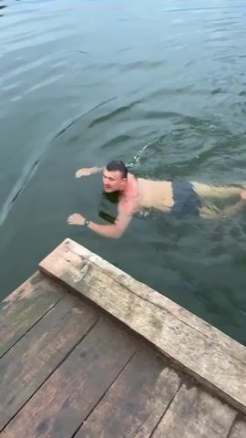 A man swims in a body of water next to a wooden dock. He is wearing black swim trunks. The water appears murky and there are ripples on the surface. In the distance, a white boat is visible. The sky is overcast.