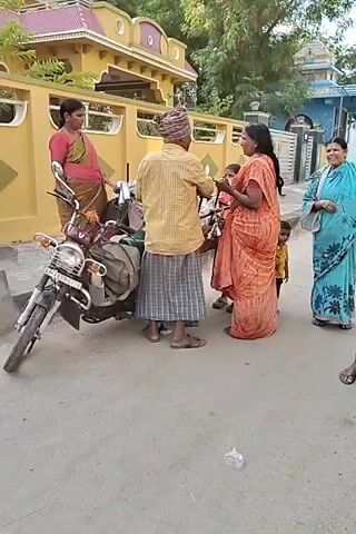 The video shows a group of people, likely in India, gathered around a cart filled with small brass religious items. A man in a yellow plaid shirt and a plaid skirt is examining one of the items, while a woman in an orange sari holds another. The cart is overflowing with similar items, some still wrapped in plastic. A motorcycle is parked nearby, loaded with luggage. Another woman in a green and pink sari stands near the motorcycle. Children are visible in the background. The scene takes place on a paved area in front of houses.