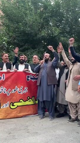 A group of approximately ten men stand behind a red and orange banner, protesting. The banner has writing in Urdu on it. The man in the center, wearing a blue shalwar kameez and a gray vest, speaks into a microphone, raising his right fist in the air. Several other men raise their hands in the air. The protest takes place outdoors near a street, with buildings and trees in the background.