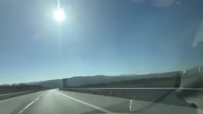 The video shows a view from inside a car driving on a highway on a bright, sunny day. The road is straight and has white lines marking the lanes. There are metal barriers on the sides of the road. The landscape in the background includes hills or mountains and some sparse vegetation. A green road sign with white lettering, indicating directions to various locations like "Kulata" and "Dupnitsa," is visible on the right side of the road. The sky is clear blue, and the sun is bright, creating a glare. There are no other cars visible in the immediate vicinity.