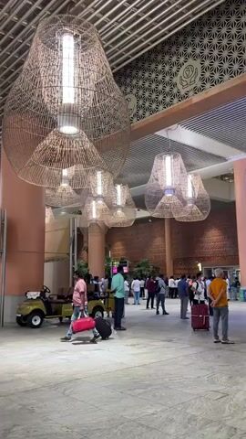 The video displays an interior view of a bustling airport terminal, panning slowly from left to right. Large, pear-shaped light fixtures made of woven, light-colored material hang prominently from a high, slatted ceiling, emitting a bright white light. The floor is light-toned and tiled. Numerous individuals, mostly adults, are present; many are walking across the floor, some pulling rolling suitcases or carrying bags. One man in a pink shirt walks from left to right, pulling a black suitcase and carrying a red bag. A yellow electric buggy, carrying two people, is seen driving from left to right. In the background, various shops, food stalls with signage (including "MITTI CAFE"), and digital information screens are visible. A large, intricate white sculptural art piece is briefly visible on the far left of the frame. The overall lighting suggests an interior night or evening setting.