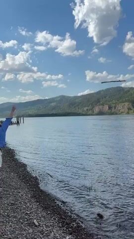 The video shows a body of water with mountains in the background and scattered clouds in the sky. The camera then pans over the water, which appears to be a river or lake, towards a shoreline where a wooden pier and some logs are visible. The camera then focuses on a young woman standing on the shore. She is wearing glasses and a blue shirt, and she is looking directly at the camera with a smile. She also wears a necklace with a cross pendant.