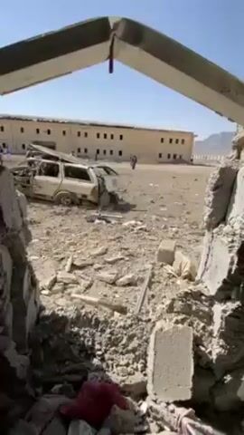 The video shows the aftermath of an explosion. A destroyed white station wagon sits in a debris-filled area outside a multi-story beige building. The camera's perspective is from inside a damaged structure, looking out through a large hole in the wall, revealing rubble and twisted metal. Several people are visible in the background near the building. The sky is clear and blue.