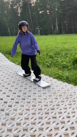 A person wearing a purple long-sleeved shirt, black pants, black gloves, and a black helmet is snowboarding. The person is on a white snowboard with a marbled design. The person is riding on a textured, grey surface. The person then rides onto a black cylindrical object. The person continues to ride on the black cylinder and then rides onto a blue mat. The person is in a grassy outdoor area with trees and buildings in the background. The sky is overcast.