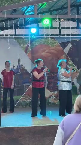 The video shows a group of three women performing on an outdoor stage in Kukui, Russia. The stage is covered by a roof, and string lights are hung above. The backdrop is a large banner with several images, including a depiction of a village with onion-domed churches, a large loaf of bread in a basket, daisies, and a cow. The women are all holding microphones and appear to be singing. They are dressed in brightly colored, shimmering outfits. The woman on the left wears a red top and black pants. The woman in the center wears a red top and black pants, and the woman on the right wears a silver sequined top and black pants. The lighting on the stage is colorful, with blue, green, and red lights illuminating the performers. The scene appears to be set during the daytime or early evening, as there is natural light visible. The overall mood seems festive, with the performers engaging with the audience. The presence of a cow in the backdrop suggests a rural or agricultural theme for the performance.