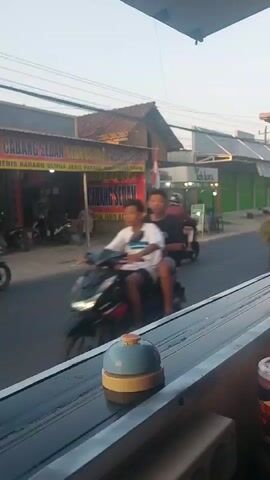 This is a vertical, handheld, video, shot from an interior perspective, looking out a window or opening. The foreground contains a blue and yellow desk bell with an orange button on top, resting on a dark surface. The background depicts a street scene. On the right side of the street, there is a shop with a red and yellow banner that reads "CABANG SEDAN" in large text and "SEMUA JENIS BARANG" in smaller text. There are motorcycles parked in front of the shop. Several people on motorcycles are traveling down the street. One person is riding a motorcycle and carrying a large bundle of green foliage. Another motorcycle has two people riding on it. A third motorcycle has three people riding on it, two in the front and one in the back. A fourth motorcycle has two people riding on it. A fifth motorcycle is visible further down the street. On the right side of the street, further down, there is a food stall with a green awning. The sky is a pale, overcast color. The overall impression is a street scene in a Southeast Asian country, possibly Indonesia, based on the text on the banner.
