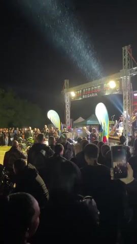 At night, a crowd of people, mostly men, gather in an open space in front of a stage. The stage is illuminated by bright lights. A sign that reads "BULGARIA" hangs above the stage. Two large teardrop-shaped flags flank the stage. Some tents can be seen behind the stage. A few motorcycles are parked near the front of the crowd. What appears to be rain or snow is falling. The scene is captured from behind the crowd.