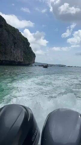 A video shows a view from the back of a boat moving through the ocean. Two boat motors are visible in the foreground. The water is choppy and creates a white wake behind the boat. In the distance, there is a large cliff face covered in green vegetation. Several long-tail boats are scattered across the water. The sky is blue with fluffy white clouds.