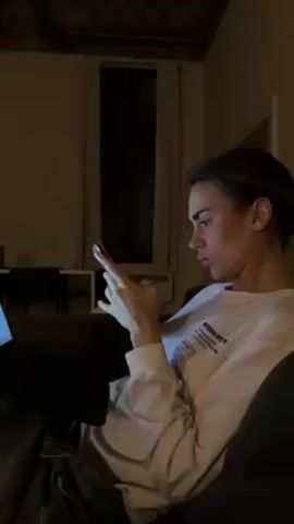 A person with light brown hair pulled back into a bun sits on a dark couch, facing left, and looking at a mobile phone in their hands. They are wearing a white long-sleeve shirt with black lettering on the front.  A black laptop sits on the person's lap, its screen illuminated. The room is dimly lit, with a window visible in the background, though it is dark outside. The ceiling has some decorative elements. A table and chairs are partially visible in the background, along with a door frame.