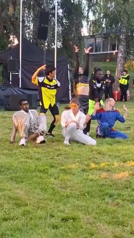 Six individuals are positioned on a grassy outdoor area in front of a stage setup with black fabric backdrop and speakers. The person on the far left, wearing a silver tracksuit, is in a low crouch with their left hand on the ground and their right arm extended. To their right, a person in a yellow and black jersey and black shorts is in a lunging pose. In the center, a person in a white jumpsuit is kneeling, touching their forehead. To the right of the person in white, another individual is kneeling and reaching out with their right arm. In the back, a person in a black top and neon green shorts is standing with their arms outstretched. To the far right, a person in a blue jumpsuit with red accents is on their hands and knees. Several other individuals are visible in the background, some wearing yellow vests, and there is a building with windows in the distance. The lighting suggests it is late afternoon or early evening.