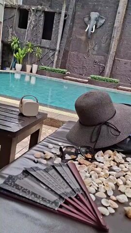 This is a short video showing a poolside scene in Sihanoukville, Cambodia. The main subjects are a wide-brimmed straw hat, sunglasses, an open hand fan with Chinese characters, and a pile of seashells scattered on a lounge chair. A small, light-colored portable speaker sits on a wooden table next to the chair. In the background, a swimming pool with clear turquoise water is visible. The pool is bordered by a wall decorated with large, sculpted elephant heads. The wall also features a textured, rock-like design and some potted plants.

The video appears to be filmed during the daytime, likely with bright sunlight given the vibrant pool water and the presence of sunglasses. The overall mood is relaxed and suggests a holiday or vacation setting, perhaps a resort. The elephant sculptures and the lush greenery hint at a tropical location, consistent with Sihanoukville. There is no explicit activity taking place other than the static arrangement of objects, creating a still-life scene.

The hand fan clearly displays Chinese characters in black ink on a dark background. The text on the fan is not clearly legible due to the angle and resolution. The seashells are of various sizes and shapes, adding to the beach-like aesthetic. The straw hat is a dark grey color with a black ribbon tied in a bow. The sunglasses are dark and stylish. The overall impression is one of serene relaxation, possibly before or after enjoying the pool.