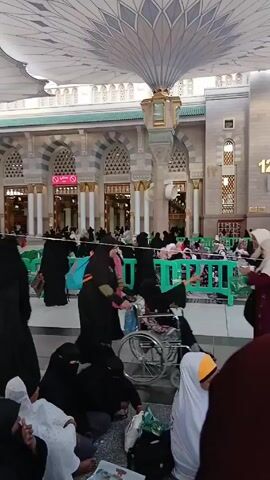 The video shows a public area with many people in traditional Islamic clothing. Many women are wearing hijabs and abayas. Some are seated on the floor, others are walking. A few are pushing a wheelchair. The background features an ornate building with arched windows and columns. A large decorative awning covers the area. Green plastic barriers are placed in the space. A sign with the number "12" is visible on the building.