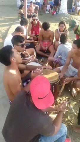 A group of young people, appearing to be teenagers or young adults, are sitting on a sandy beach. Some are wearing swimwear, while others are in casual clothing. In the center of the group, two individuals are playing drums, specifically conga drums. One is using drumsticks, while the other is playing with their hands. Other members of the group are watching, some with drinks in hand. In the background, more people are visible further up the beach, some sitting and others standing. The environment appears to be a sunny day, likely at a beach resort or popular gathering spot. The overall atmosphere seems relaxed and social.