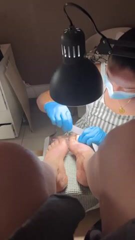 The media file captures a close-up view of a person's feet receiving a pedicure. A manicurist, wearing blue gloves and a face mask, is carefully filing one of the toenails under the bright light of a task lamp. The setting appears to be a nail salon or a dedicated space for pedicures, indicated by the presence of a foot spa device and the manicurist's professional attire. The lighting suggests it could be daytime, though the interior setting makes it hard to determine the time of day or weather. The focus is on the meticulous process of nail care, with the manicurist concentrating on her task. The perspective is from the client's viewpoint, looking down at their own feet and the manicurist at work. There is no visible text in the media file.