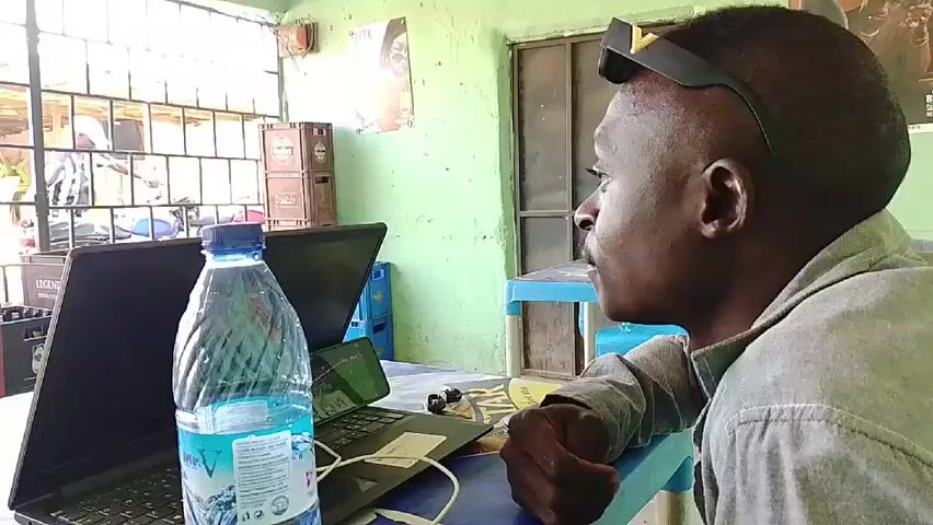 A man is sitting at a table indoors, facing right. He has short hair and is wearing sunglasses on top of his head and a light gray shirt. He is looking at something out of frame. On the table in front of him are a laptop, a phone, a bottle of water, and other small objects. Behind him, the wall is light green and there is a window to the left. A stack of crates is visible through the window. There is also a door behind him to the right and some posters on the wall.