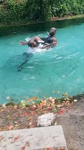 In an outdoor setting, two dark-skinned men are in a clear, blue-green body of water. The water's bottom is slightly visible, scattered with debris. One man is floating on his back, with the other man positioned vertically in the water, facing the floating man. The vertical man appears to be holding the floating man, who is resting his legs on the other man's shoulders, with his arms raised. Lush green vegetation and trees line the water's edge in the background. The foreground features a concrete slab and a strip of ground covered with fallen leaves and small debris. The overall lighting is bright and sunny.