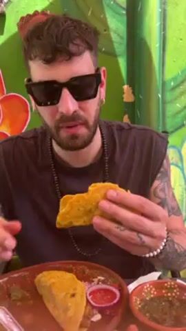 A fair-skinned man with tattoos, wearing sunglasses and a black sleeveless shirt, is eating a yellow taco. He has a beaded necklace and a white beaded bracelet. He is seated at a table with a brown plate that has another taco and a small dish with red onions and salsa. There is also a small bowl of green salsa. The background is a brightly colored green wall with a large orange flower painted on it. The man takes a bite of the taco and seems to enjoy it.