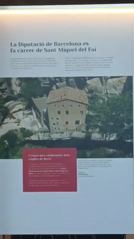 The video shows a close-up, slightly tilted view of an informational display. The display features a large photograph of a stone building nestled in a rocky, wooded landscape. Text is present above and below the photograph. The text above the photo appears to be a title and descriptive paragraphs in Catalan. Below the photo, a red square contains more text, also in Catalan, with some smaller text in the lower right corner of the display, potentially credits or additional information. The overall lighting suggests the display is indoors, possibly in a museum or exhibition space. There are no people or direct interactions visible in the frame.