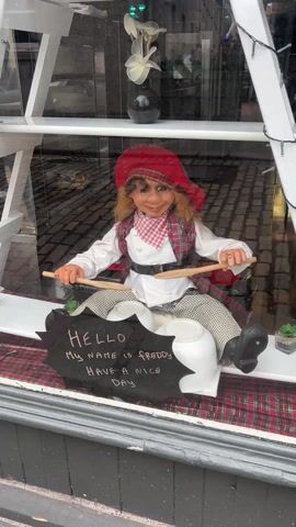 The video shows a window display. Inside the window is a mannequin doll dressed in a white shirt, plaid vest and cap, and checkered pants. The doll is sitting and holding two wooden spoons, as if playing drums on two white pots. A sign in front of the doll says, "Hello, My name is Freddy, Have a nice day." The display is on a checkered cloth, and there is a small potted plant. The window is framed by white shelves and string lights. Cobblestone pavement is visible through the glass.