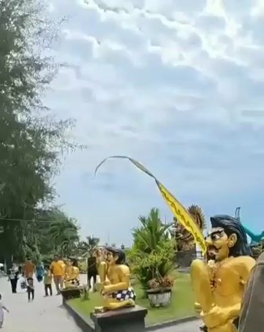 The video shows a sunny outdoor scene with a cloudy sky. In the foreground, a large golden statue of a character with dark hair and a mustache is prominently displayed. The statue is sitting and appears to be holding an object. Behind it, a long yellow banner with writing on it is attached to a pole and flutters in the wind. To the left of the golden statue, there are several smaller golden statues of people in traditional checkered cloths. People are walking around in the background, some appearing to be tourists. There are trees and green foliage in the environment, suggesting a park or public space. Further in the background, there are other colorful statues, including one of a mermaid and a white bird, indicating a decorative or amusement area. The overall atmosphere seems casual and lively.
