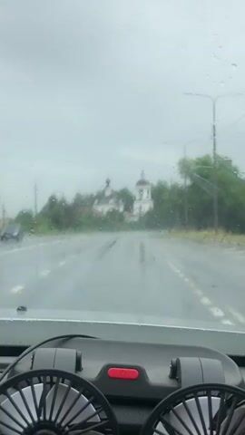 The video is filmed from inside a car looking out through the windshield. The windshield wipers are intermittently clearing the view, indicating it is raining. The road ahead is wet and appears to be a highway or a large paved road with lane markings. In the distance, there are two white church-like buildings with domes visible through the rain and trees. The sky is overcast and gray. The interior of the car is partially visible in the foreground, showing a dashboard with two black fans and a red button. A car with its headlights on is visible on the right side of the road in the distance.