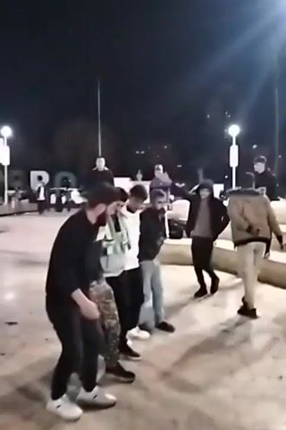 At night, a group of about nine men are dancing in a public space. Most are wearing casual clothes and seem to be enjoying themselves. Some have beards. There are streetlights and a large decorative sign in the background, along with other people walking and cycling through the area. The surface they are dancing on appears to be concrete.