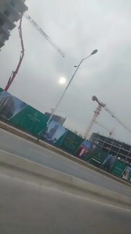 The video shows a scene captured from inside a moving car, possibly the passenger seat. The camera is angled towards the right side of the road. Outside, there is a view of a construction site with several tower cranes visible against a cloudy sky. A partially constructed building frame can be seen behind a barrier with promotional advertisements on it. A grey Chevrolet Spark with license plate 30 R 451 NB is driving ahead of the cameraman. The video shifts to show the driver of the car, seen from the back, a man with short dark hair wearing a dark colored sweater. He is focused on the road ahead. The overall impression is of a dull, overcast day at a construction site, as seen from inside a moving vehicle.