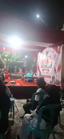 The video shows an outdoor gathering at night. A stage is set up with a red carpet and a backdrop featuring an Indonesian flag and text that reads "REPUBLIK INDONESIA 1945-2026". Several people are on the stage, some seated and some standing. One person in a white shirt appears to be playing a game or interacting with a table. In the foreground, an audience is seated on chairs, facing the stage. Red and white balloons are strung up on the left side of the frame. Lighting from the stage illuminates the scene.