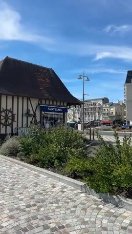 The video pans across a street scene on a sunny day. In the foreground, a cobblestone walkway is visible, bordered by low green bushes. To the left, a building with a dark tiled roof and half-timbered accents features a blue awning with the words "SAINT JAMES" above a storefront. Further down the street, several multi-story buildings with light-colored facades and dark roofs are visible, some with balconies. On the right, a larger, older building with a prominent dark tiled roof and half-timbered detailing stands out. Cars are parked along the street and some are in motion. A few people are seen walking on the sidewalk in front of the buildings. The sky is bright blue with scattered white clouds.