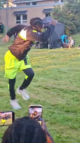 A Black man with dreadlocks and yellow sunglasses is performing a dance routine outdoors on a grassy area. He is wearing a black athletic jacket with yellow accents, bright neon green shorts over black leggings, and white athletic shoes. His body is in a low, wide stance, with his hands clasped in front of him. In the background, there is a stage with lighting equipment, a tent, and several people observing the performance, some of whom are also dancing. Two mobile phones are visible in the foreground, held by unseen individuals, recording the dancer. The setting appears to be a park or an outdoor event space, with trees and a building visible in the distance. The lighting suggests it is daytime or early evening.