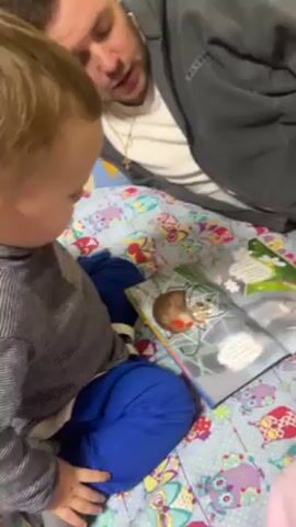 Here's a description of the video:

A man is lying on a bed and reading a book to a toddler. The man is wearing a grey jacket over a white t-shirt and has a cross necklace. The toddler is wearing a striped shirt and blue pants. They are both looking at the book, which has a picture of a spider on one page and other illustrations on the opposite page. The bed is covered with a colorful patterned blanket.