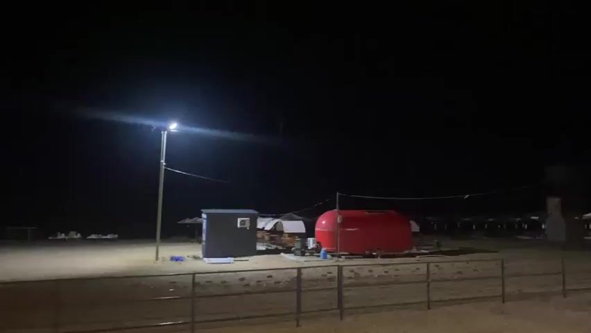 This is a video of an outdoor scene at night, illuminated by a single streetlamp. The ground appears to be sand or dirt. There is a small, dark blue utility box with a window or panel on the front. To the right of the utility box is a large, bright red cylindrical tank. In the background, several white structures with pointed roofs are visible, possibly tents or small buildings. There are also some fence-like structures in the distance. The streetlamp is the primary light source, casting a bright pool of light on the ground. The sky is completely dark. There are no people visible in the video.