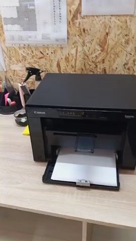 The video shows a black Canon printer on a wooden desk. A sheet of white paper is in the printer's output tray. The printer has a black exterior and appears to be in a home or office setting. The background consists of a wall made of wood panels, with some papers and office supplies visible on the wall and on the desk. The printer's power light is on. There are no people visible in the video.