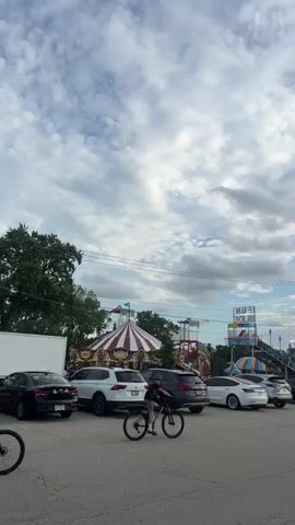 The video shows a parking lot adjacent to a carnival. In the foreground, two people are riding bicycles. In the background, various carnival rides are visible, including a carousel and a slide. Cars are parked in the lot. The sky is cloudy.