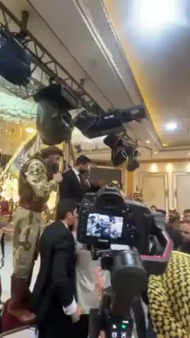 The video shows a lively scene inside what appears to be a wedding reception hall. A group of men are gathered around a stage or slightly elevated area, some are dressed in traditional middle eastern attire, while others are in suits. One man, dressed in camouflage clothing and boots, is standing on a higher level, microphone in hand. He appears to be addressing the crowd.  Another man in a suit also has a microphone and seems to be speaking. The other men in the crowd are cheering and raising their hands. Several lights are visible above them, and a professional camera on a tripod is recording the event from the foreground. The overall atmosphere seems festive and celebratory.