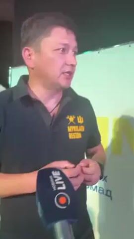 A man is being interviewed. He is wearing a black polo shirt with the words "MYKOLAIV REGION" printed in yellow on the left breast. The emblem of Ukraine is also printed on the shirt. The man is standing in front of a white and yellow backdrop. A microphone with the words "LIVE" and the logo of the "Q" Ukrainian television channel is held up to him.  Another person wearing a headdress is visible in the background on the left side of the screen.