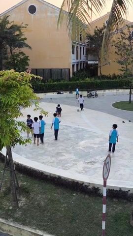 Several young boys are playing soccer on a concrete surface.  One of the boys falls to the ground, and another is kicking a soccer ball. One boy is standing apart. Another boy drives by in the background on a motor scooter. A tree is on the left side of the frame and there is a building in the background. There is a pedestrian prohibition sign on the right side of the frame.