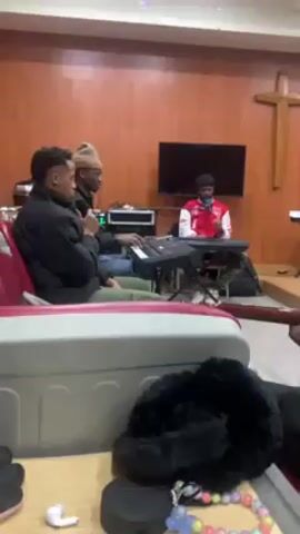 The video shows a group of people in a room that seems to be a church, given the wooden cross on the wall. There are four people visible, and they are all involved in playing music. Three of them are sitting, and one is standing. Two people are playing keyboards, one is singing into a microphone, and the fourth person is playing the guitar. There is a large TV screen on the wall behind them. There are also other objects in the foreground, including a black fluffy item, some headphones, and a colorful beaded necklace. The walls are covered in wooden panels, and the overall lighting in the room is warm. The camera is positioned low and to the side, providing a view of the group from a slightly obscured angle.