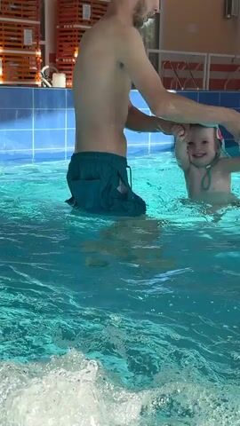 A man in swim trunks and a swim cap is in a swimming pool, holding a child's hand. The child is wearing a swim cap with a colorful bird design and appears to be being assisted in the water. The pool has blue tiled walls and some orange shelving units are visible in the background, along with colorful triangular flags. The water is clear and blue, with ripples and splashes indicating movement. The man is facing the child and appears to be instructing or supporting them. The child is looking down and moving their legs in the water.