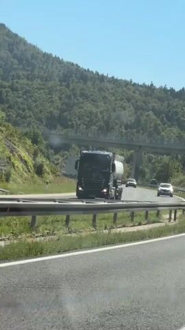 A black truck with a white cylindrical tank is driving on a highway. To the right of the truck, a white sedan is also driving on the highway. The highway is surrounded by a green grassy area and lush green trees on a mountainside. A concrete overpass crosses over the highway in the distance. A speed limit sign indicating 100 is visible on the right side of the highway. The sky is clear and blue.