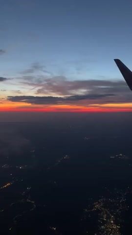 The media file is a vertical video capturing an aerial view of a sunset from an airplane. The main subject is the vast expanse of the sky and the earth below. The setting is the view from an airplane window, with a portion of the airplane's wing visible on the right side of the frame.

The activity taking place is the descent of an airplane, as suggested by the illuminated city lights visible on the dark land below, indicating it is either evening or early morning. The sky is a gradient of colors, transitioning from a deep blue at the top to shades of orange and red near the horizon, characteristic of a sunset. Wisps of clouds are scattered across the sky, catching the light of the setting sun.

Notable details include the illuminated city lights, suggesting a populated area below. The time of day is clearly sunset or twilight. The airplane wing is a prominent object, with a visible logo on its tip, though the text is illegible. The overall emotion conveyed is one of awe and beauty, inspired by the breathtaking natural phenomenon of a sunset viewed from high above the earth. The location context provided indicates this scene is over Catanzaro, Italy.