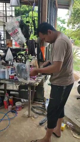 A man stands in an open-air workshop, working on a black object encased in plastic. He is wearing a brown t-shirt and black shorts. He holds a tool in his right hand, which appears to be an electric sander or grinder, and is focused on the object. The object is mounted on a metal stand. The workshop is cluttered with various items including tools, paint cans, plastic bags, and parts of motorcycles. The floor is concrete, and there is a blue hose coiled on the ground. In the background, there are motorcycles and more workshop equipment. The overall environment suggests a repair or fabrication setting.