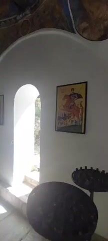 The video shows the interior of what appears to be a small church or chapel. The walls are white and adorned with several framed religious icons. One icon depicts a figure on horseback, possibly St. George. There is an arched doorway leading to an outdoor area with bright sunlight. The ceiling is decorated with religious murals and features a large, ornate gold chandelier. In the center of the room, there is a stone structure with what seems to be a small pool or fountain. The overall impression is of a small, but historically significant and well-preserved sacred space.