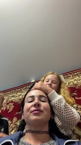 The video shows two individuals, a woman and a child, in an indoor setting. The woman is lying on a bed, holding a large knife. The child is leaning over her. The room appears to be a bedroom with a red rug on the floor and a white bed in the background. The lighting suggests it could be daytime. The woman is smiling, and the child appears to be looking at the camera. The activity depicted is unclear due to the blurry nature of the video and the close-up shots. There is no visible text in the video. The location is stated as Bronnitsy, Russia.