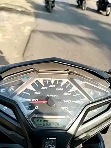 The video begins with a close-up of a motorcycle's dashboard, showing a speedometer. The camera then pans up to reveal a view from the motorcycle, showing a road with several other motorcycles and people. The sky is clear and blue, and there are buildings and trees on either side of the road. The motorcycles are all moving forward, and some of the riders appear to be waving. The road appears to be in a rural or semi-rural area. The video is brief and appears to be a point-of-view shot from a motorcycle rider.