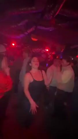 The video, recorded indoors, shows a young woman with shoulder-length brown hair, wearing a black tank top and blue jeans, dancing at a bar or club. The lighting is dim, illuminated by red and blue flashing lights, giving the room a festive yet slightly hazy ambiance. Other people are visible in the background, some dancing or simply standing, all partially obscured by the low light and motion. The camera angle is unsteady, likely held by someone moving along with the crowd, causing the view to tilt and swirl around the primary subject. The woman seems to be enjoying herself, smiling and making a peace sign towards the camera at one point. The general atmosphere suggests a lively and crowded social gathering.