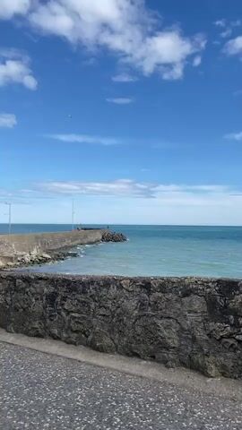 A wide shot shows a body of water and a breakwater under a partly cloudy sky. The camera is positioned at ground level behind a low, rough-hewn stone wall. The breakwater extends from the left of the frame into the distance, curving slightly to the right. It is constructed of stone blocks and appears to have lights or poles along its length. The water is a vibrant blue-green and shows small waves crashing against the breakwater. A person is visible standing on the breakwater, near the end. In the distance, a small object, possibly an airplane or a bird, is visible against the sky. The sky is a bright blue with scattered white clouds.