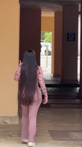 This media file captures a rear view of a person with long, dark hair walking away from the camera. They are wearing a pink, patterned long-sleeved shirt and loose-fitting lavender pants with white sneakers. The setting appears to be an outdoor walkway or corridor, with beige walls on either side. In the background, a doorway or opening reveals a glimpse of the outside, where trees and a white vehicle are visible. There is a blue sign with a white "P" symbol and an arrow pointing left, indicating parking. The lighting suggests it is daytime, and the scene seems to be taking place in Higüey, Dominican Republic. The overall impression is of someone casually walking through a building's covered walkway.