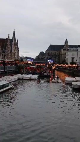 A boat navigates a canal lined with buildings and boats. On the canal's edge, there is a gathering of people in front of a stage with the words "POLE POLE" displayed. String lights illuminate the area. The sky is overcast.
