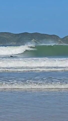 The video shows a beach on a sunny day. The water is light blue, and there are several waves crashing on the shore. In the distance, there is a hilly landscape with vegetation. A single surfer is seen paddling in the water near a larger wave. A bird flies in the clear blue sky above the scene.